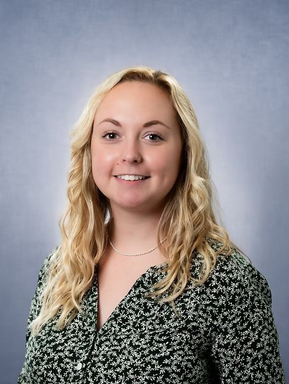 Woman with blonde hair wearing a patterned shirt smiles at the camera against a blue backdrop.