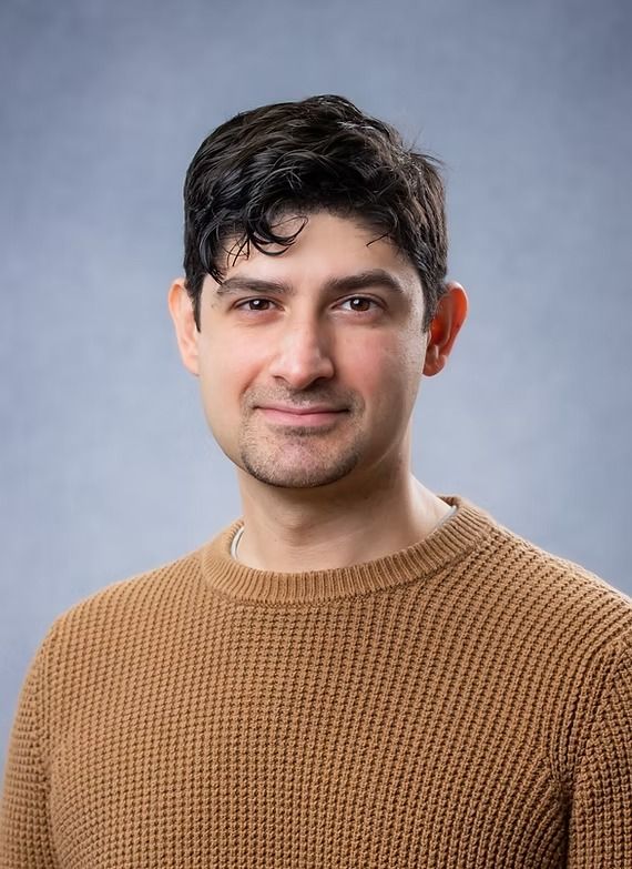 Man in a brown sweater smiles at the camera against a light blue background.