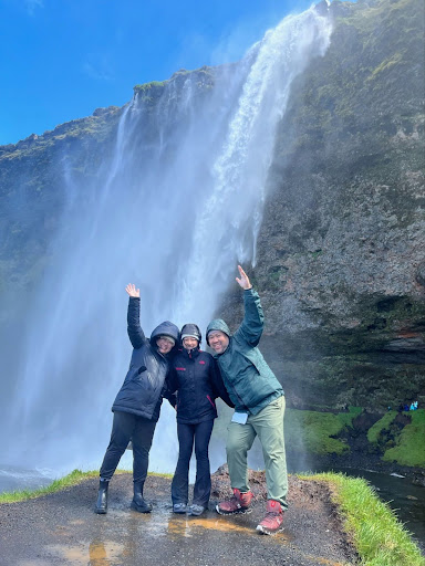 Three people wave in front of a tall waterfall with blue sky; they're wearing jackets in Iceland.