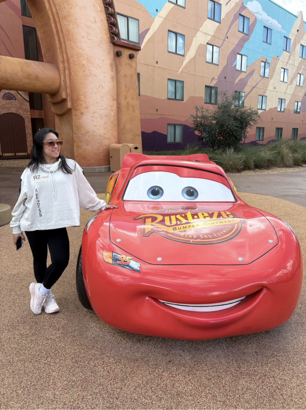 Woman posing next to a red Lightning McQueen car, in front of a building with colorful murals.