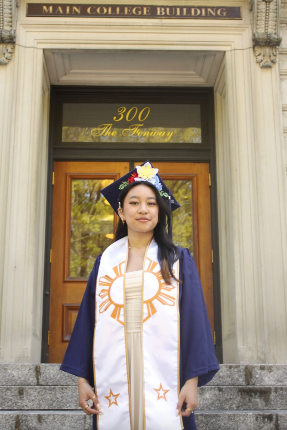 Graduating student in cap and gown smiles in front of a building with a door, wearing a sash with sun design.