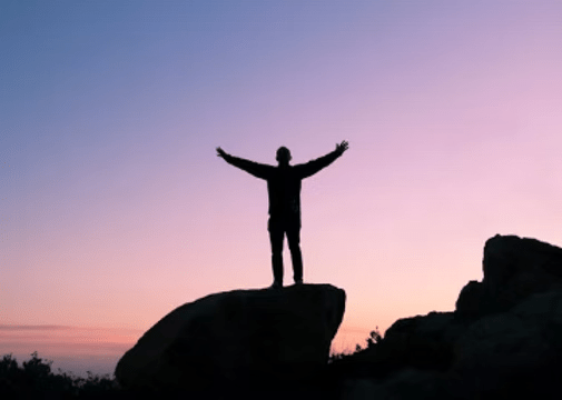 Silhouette of person with arms raised, standing on a rock at sunset. Purple and pink sky.