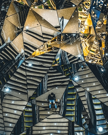 Man on escalator reflected in mirrored ceiling with geometric patterns.