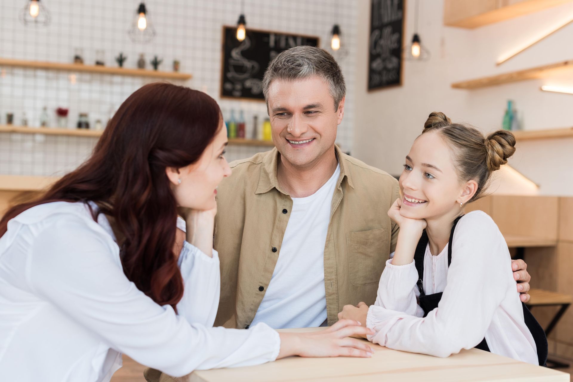Family smiling, talking at a cafe table. Man has arm around teen, woman leaning towards them.