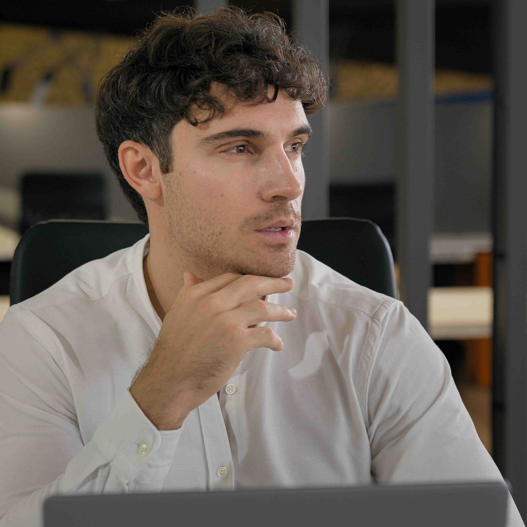 Man with curly hair in a white shirt, looking thoughtful, hand on chin, in an office setting.