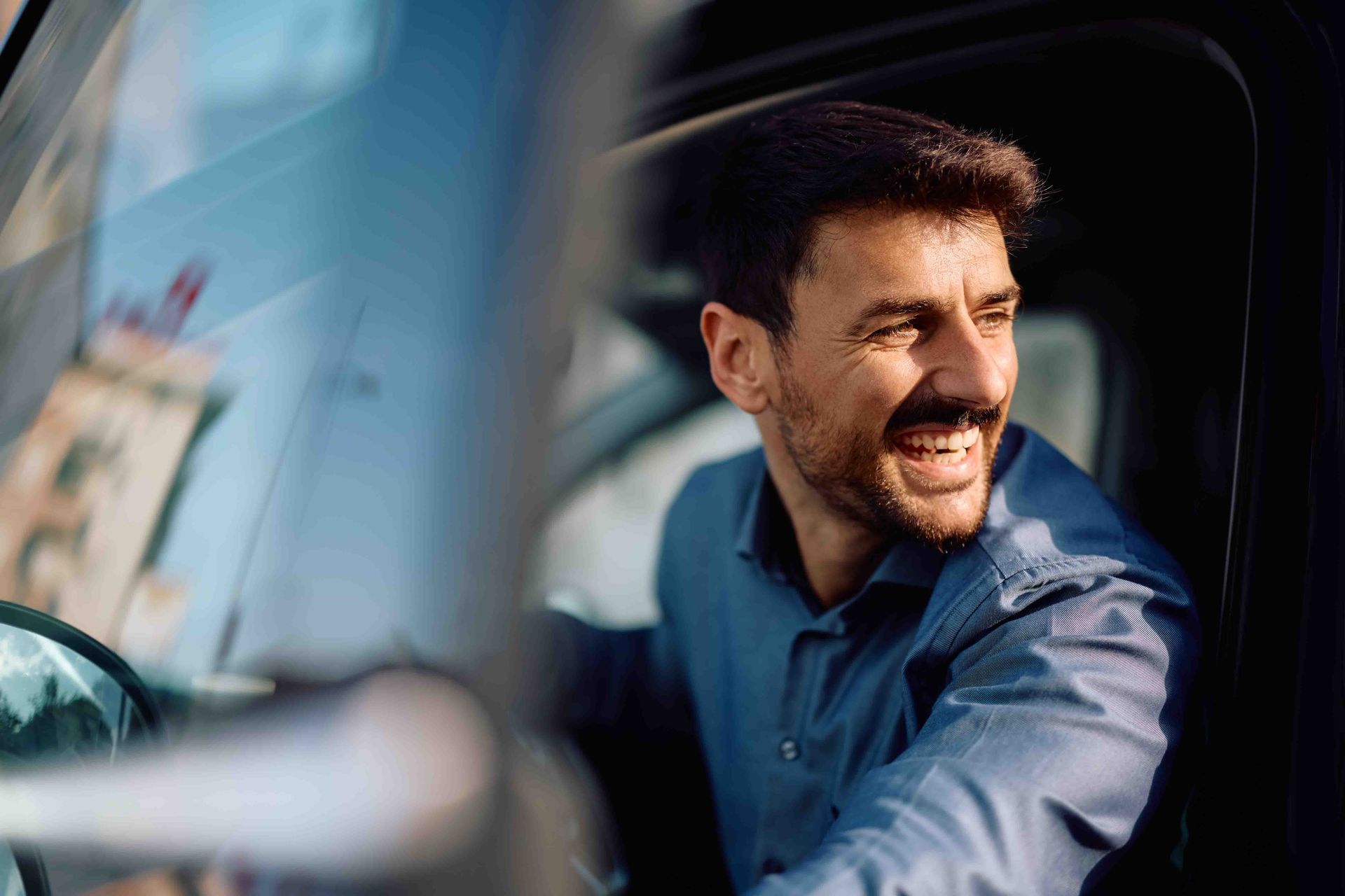 Man smiling, seated in a car, looking to the side. Wearing a blue shirt, daylit, outdoors.