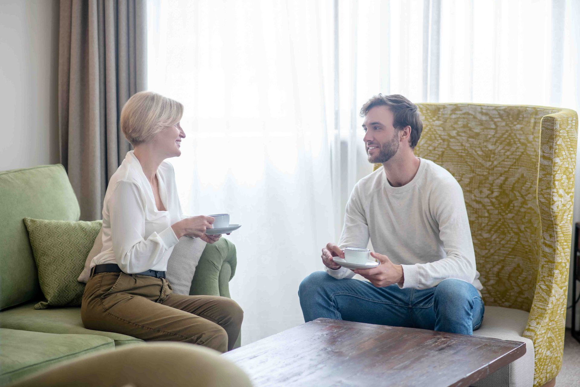 Woman and man seated, holding cups, conversing in a living room; window in background.