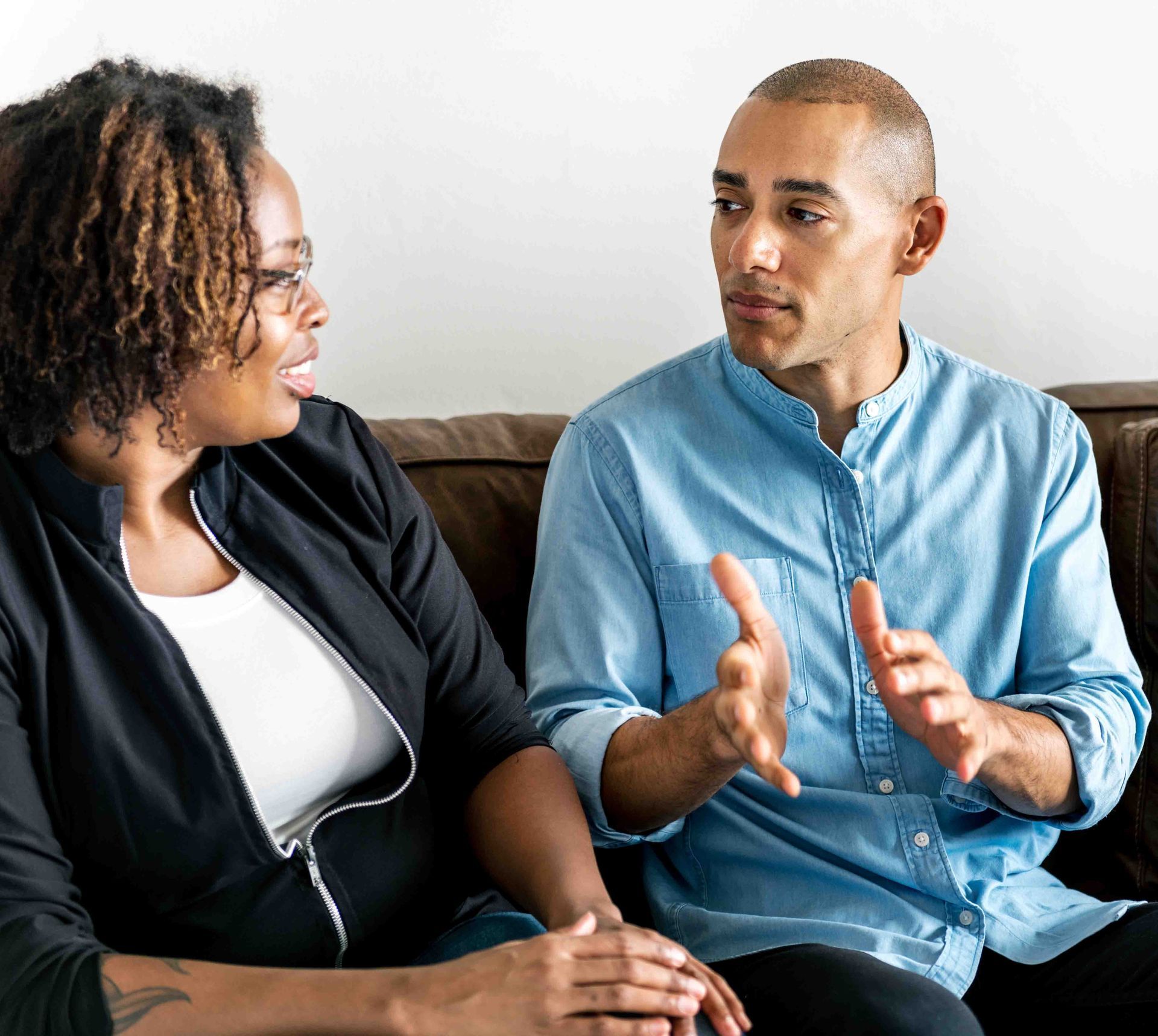 Man and woman talking, sitting on a brown couch. Man gestures with hands.