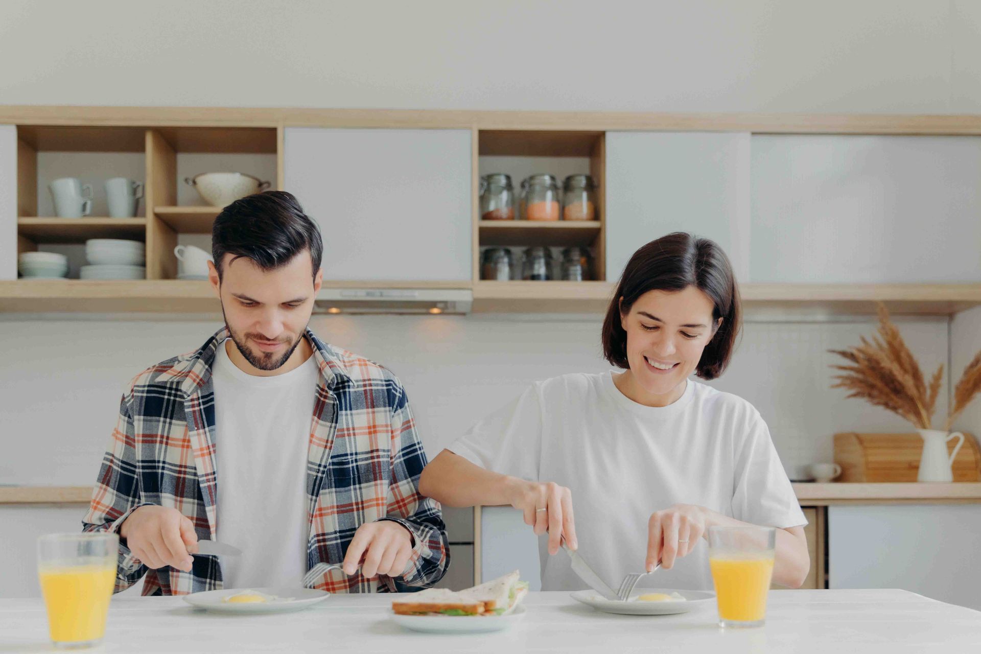 Couple eating breakfast at a kitchen table. They are smiling and cutting food with utensils. Orange juice is on the table.