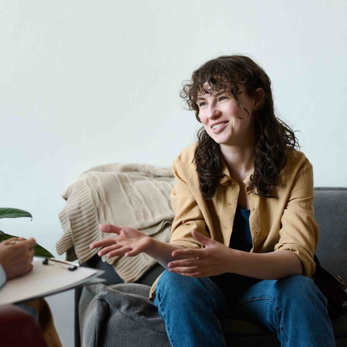 Woman smiling, gesturing, talking on a sofa, possibly at a counseling session. A person holds a clipboard.