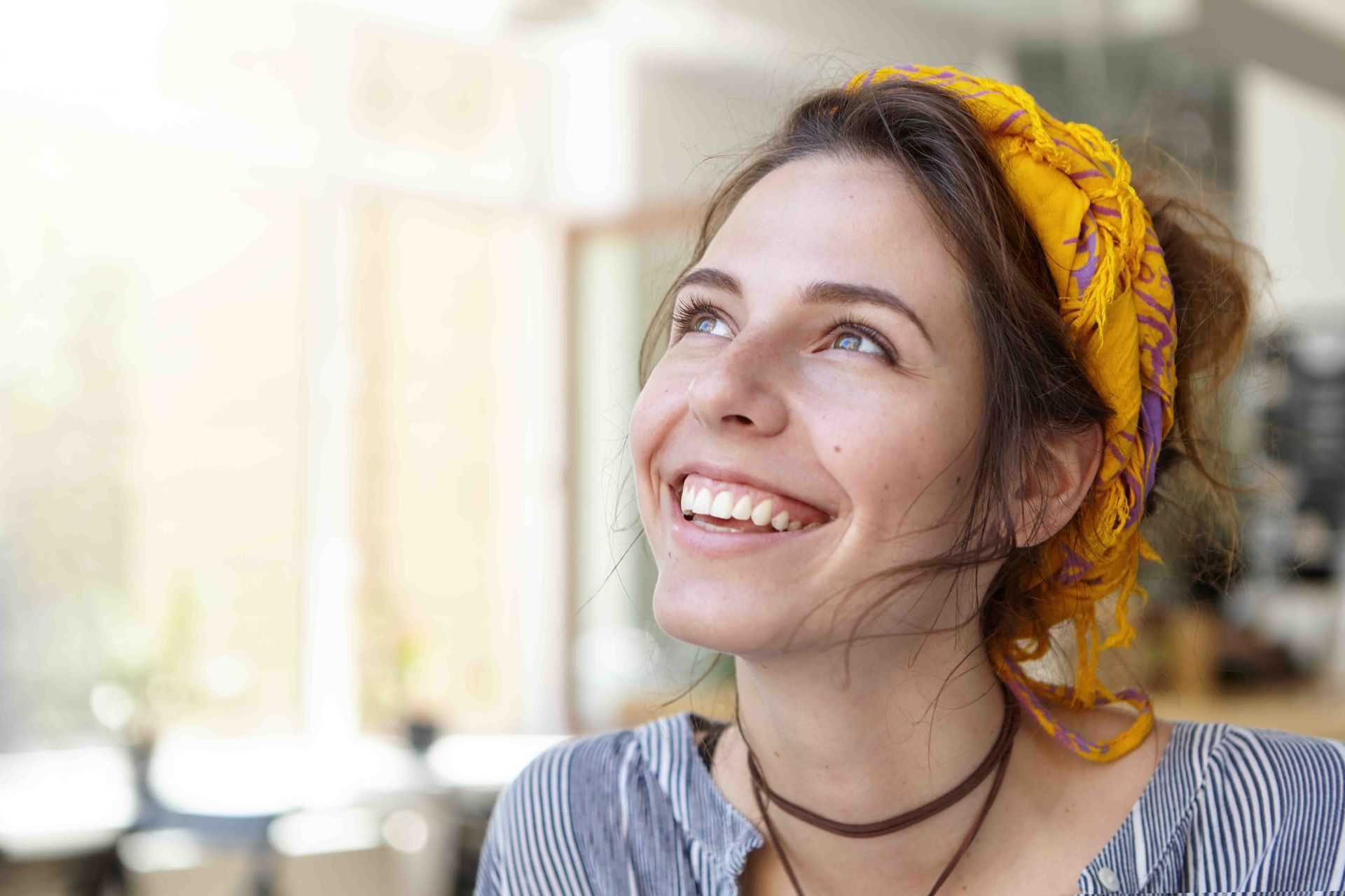 Woman smiling, looking upward. Wearing a yellow headscarf, striped shirt, and brown necklace.