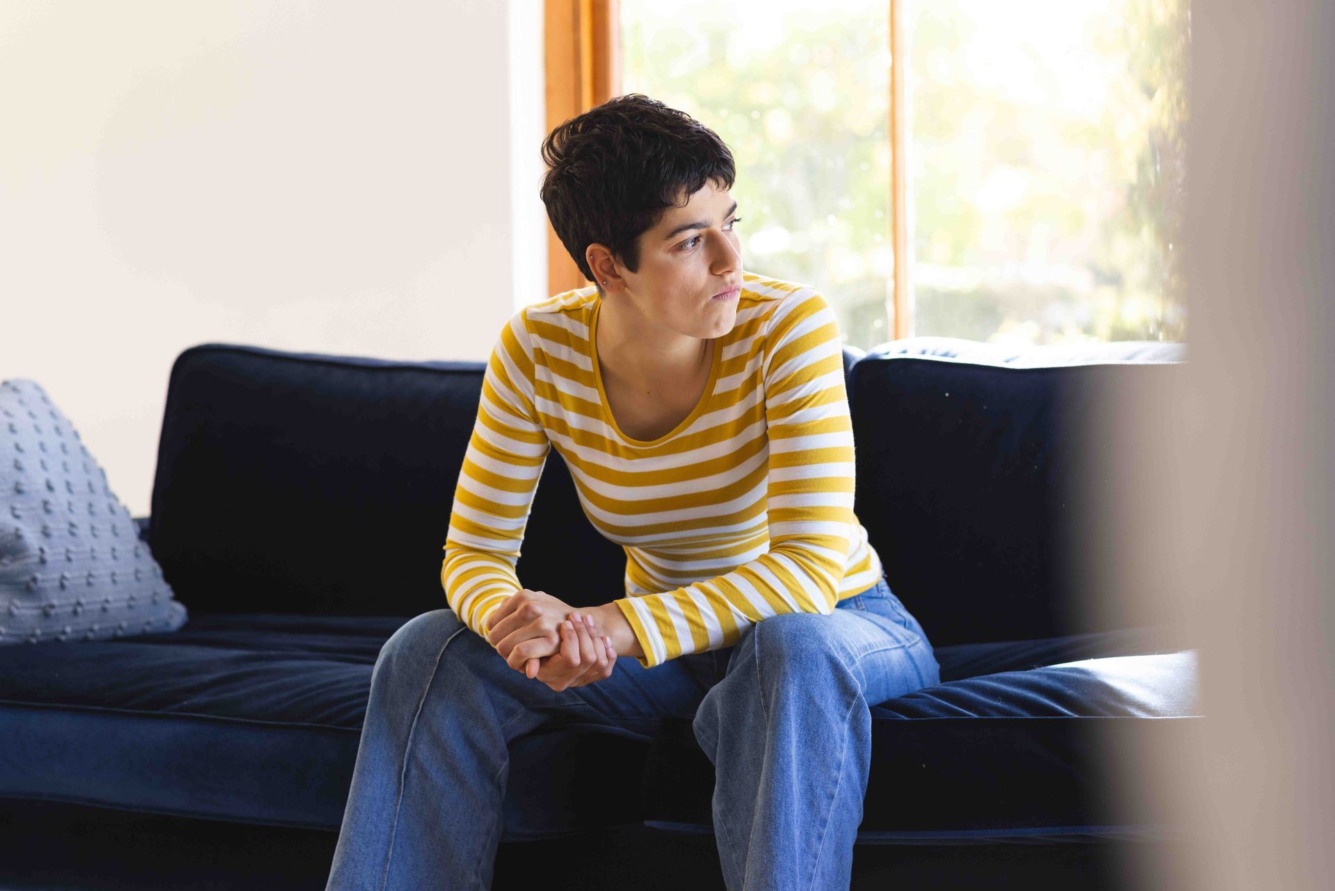 Woman sitting on a dark blue couch, gazing out a sunny window, wearing a yellow striped shirt and jeans.