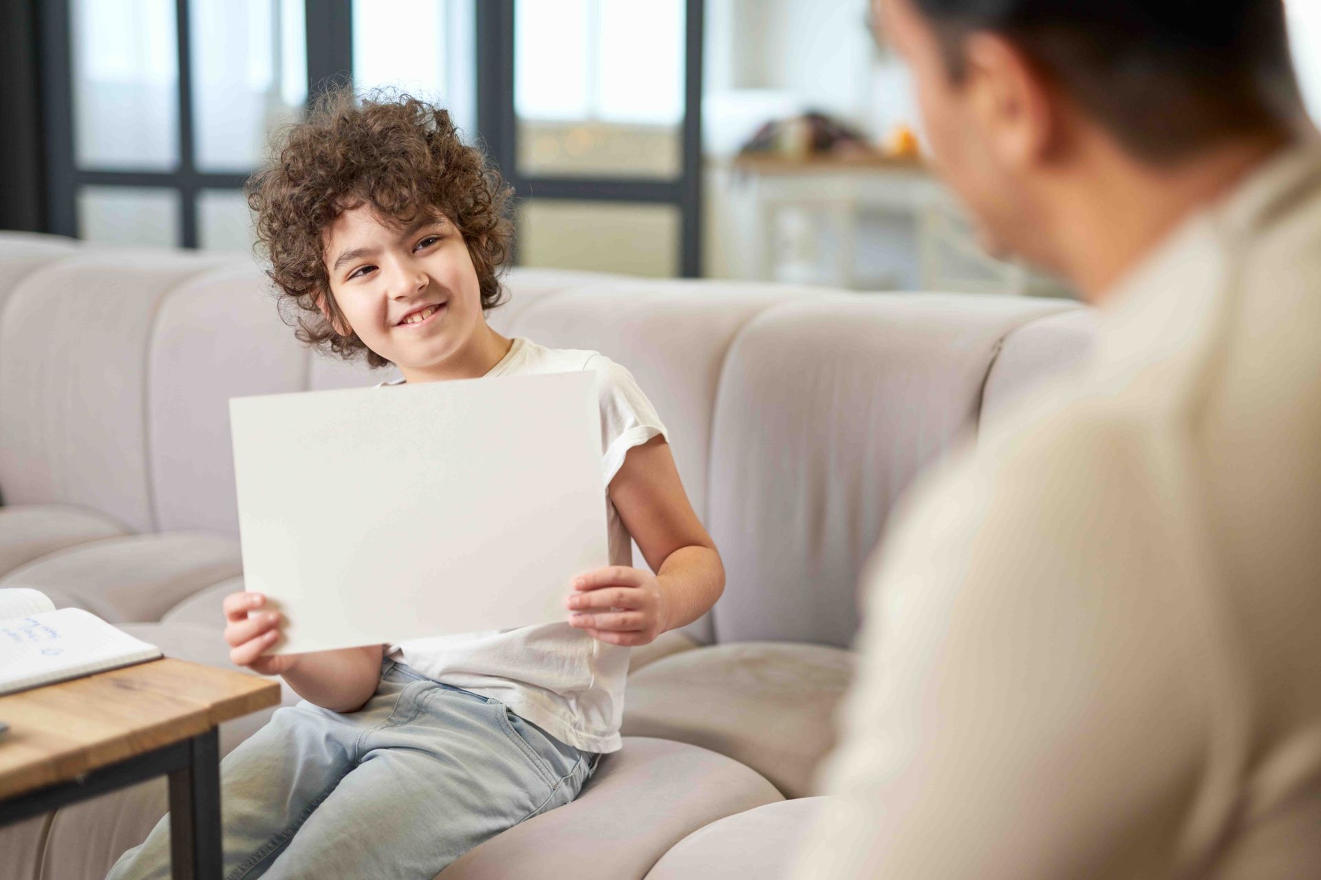 Child holding blank paper, smiling at person sitting across from them on a sofa. Living room setting.