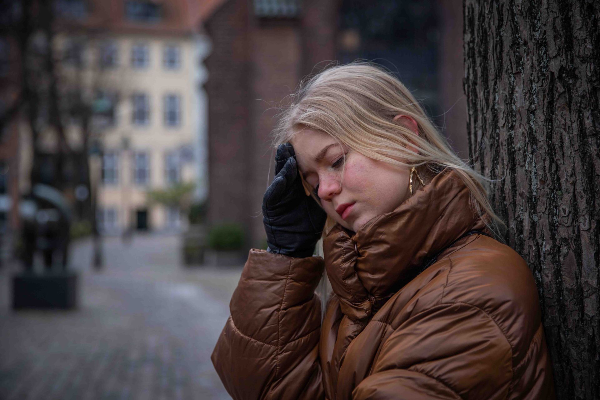 Woman leaning against a tree, eyes closed, hand on her head, wearing a brown jacket, black gloves, and earrings.