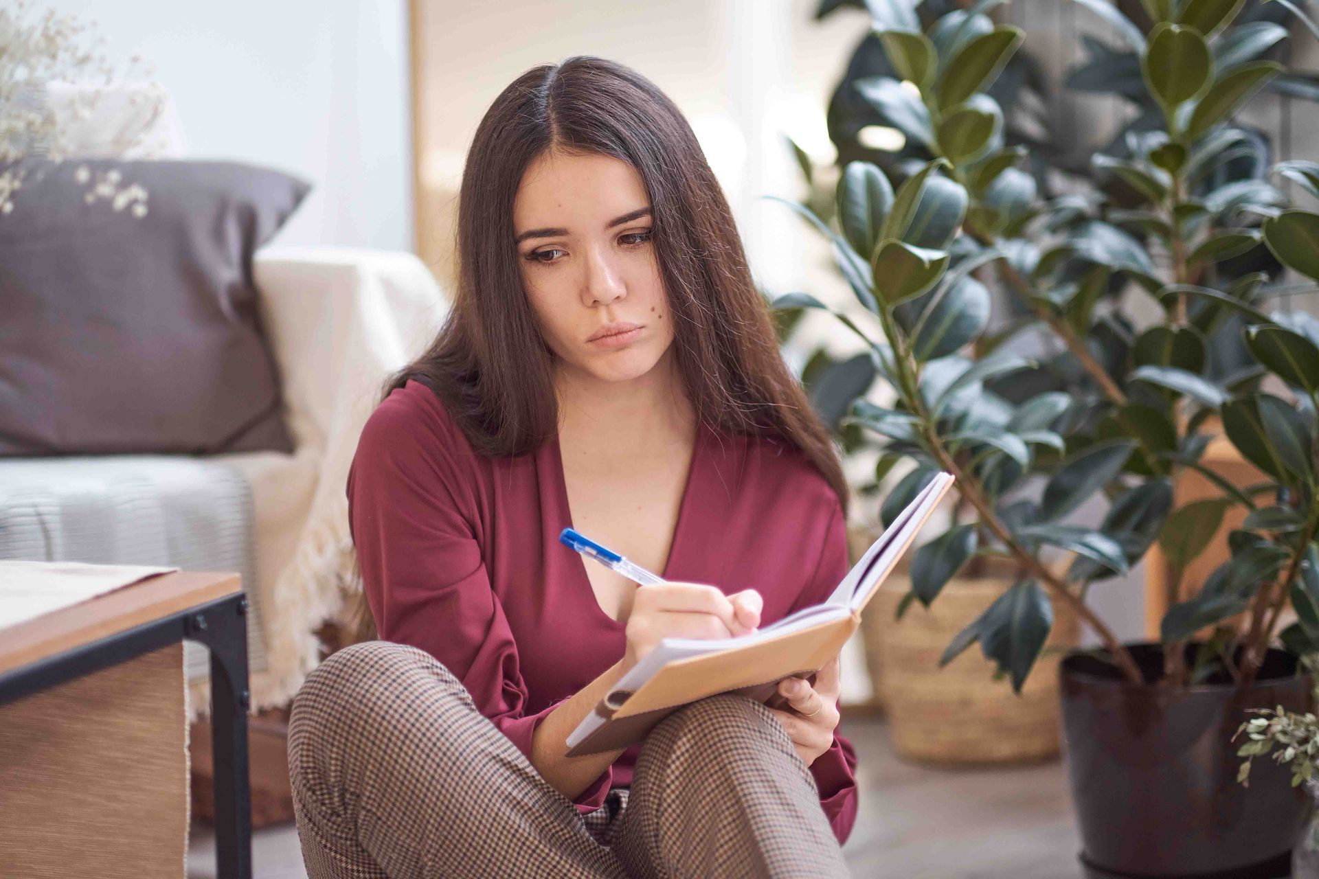 Woman sitting, writing in a notebook, looking thoughtful. Indoors, near plant and couch.