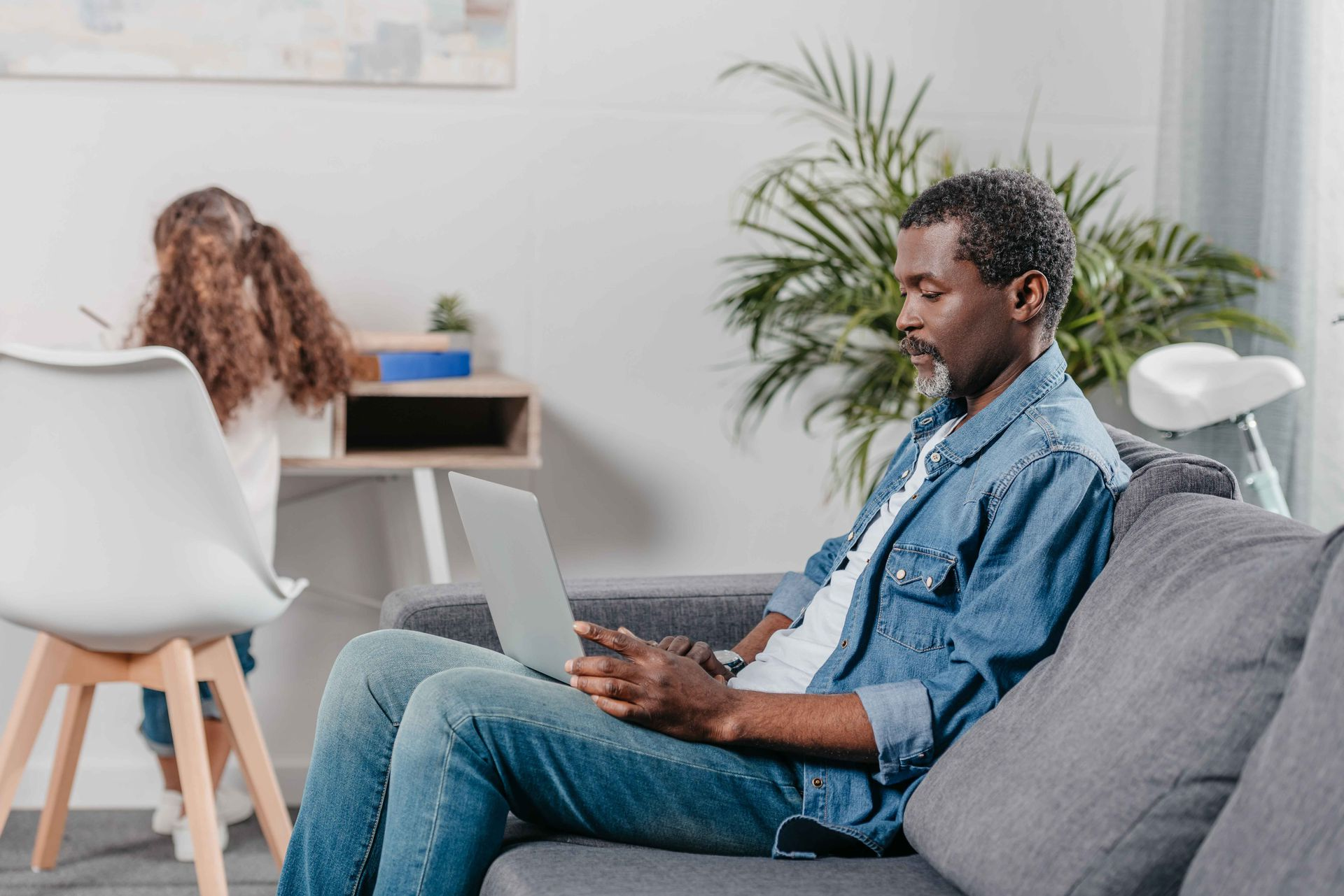 Man on couch using laptop, child near desk with paper, living room.