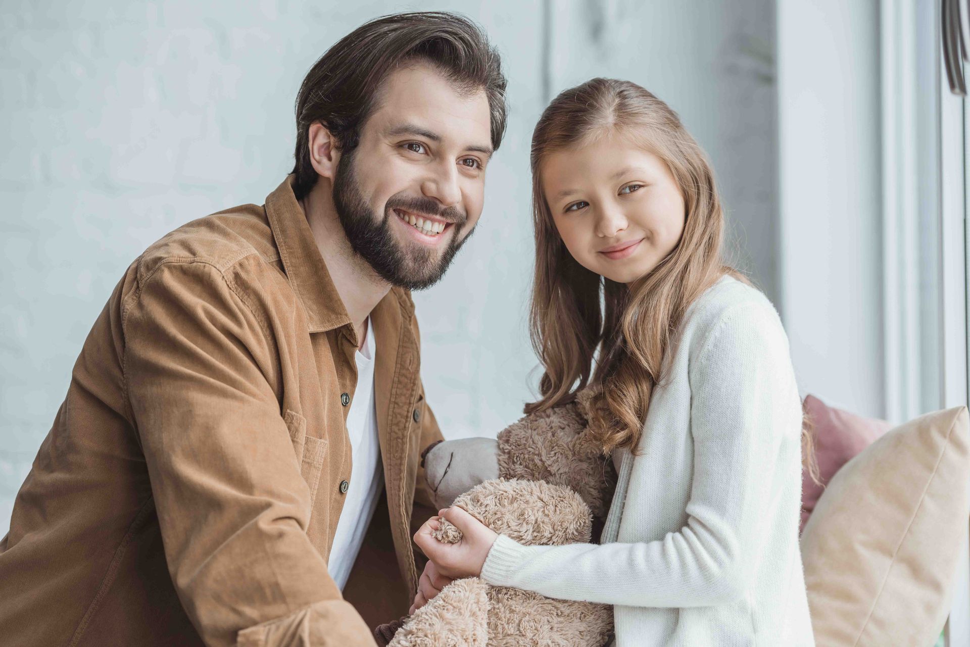 Man and girl, smiling, with a teddy bear, seated by a window.