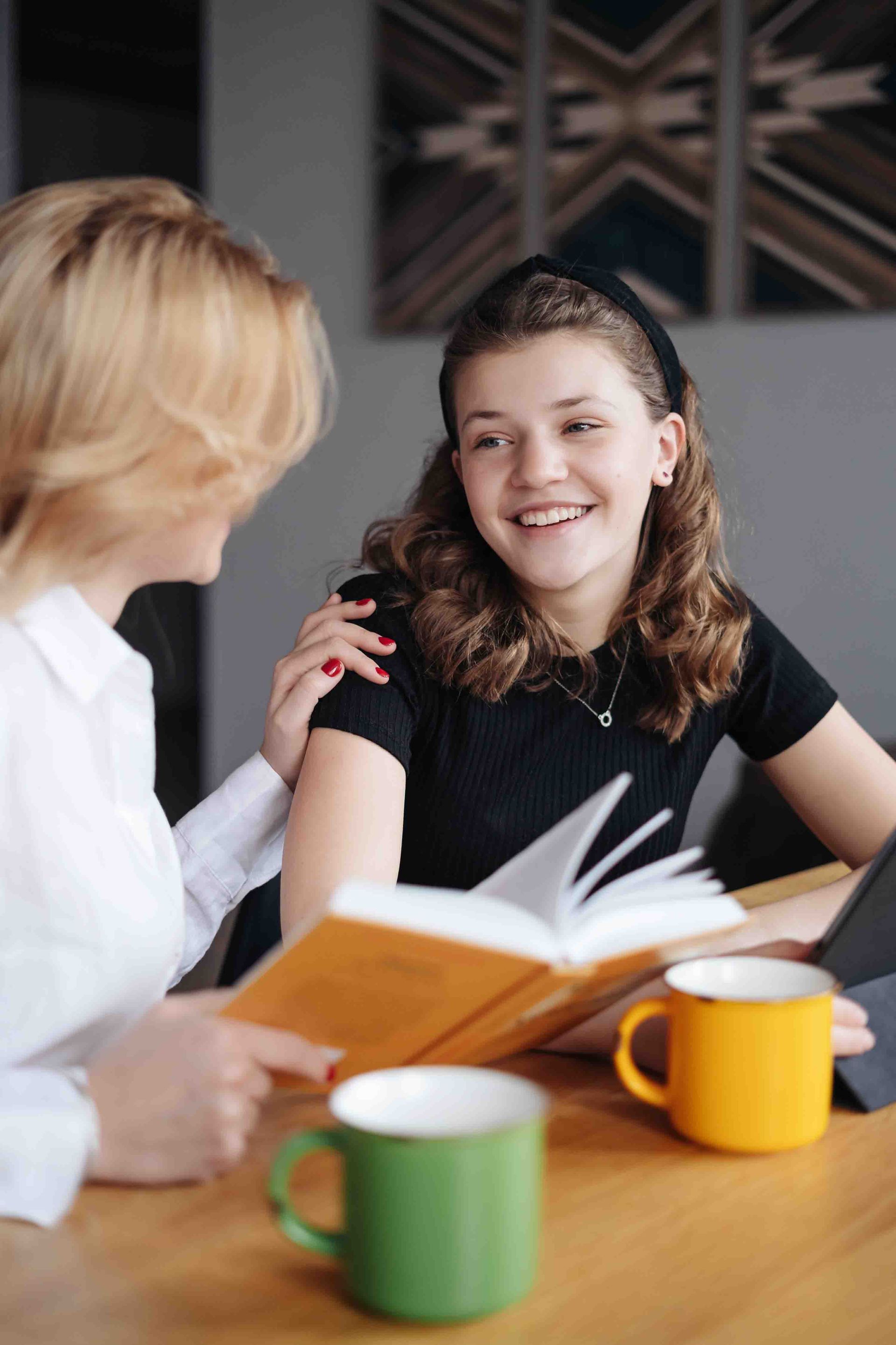 Woman points to a book while a smiling girl sits next to her, arm around her shoulder. Two mugs are on the table.