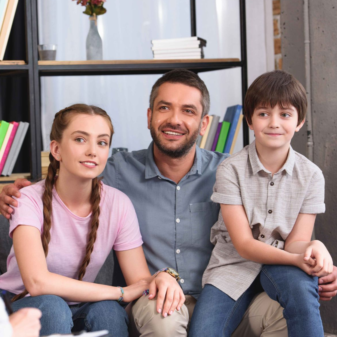 Family sitting together indoors, smiling. Father has arm around each child.