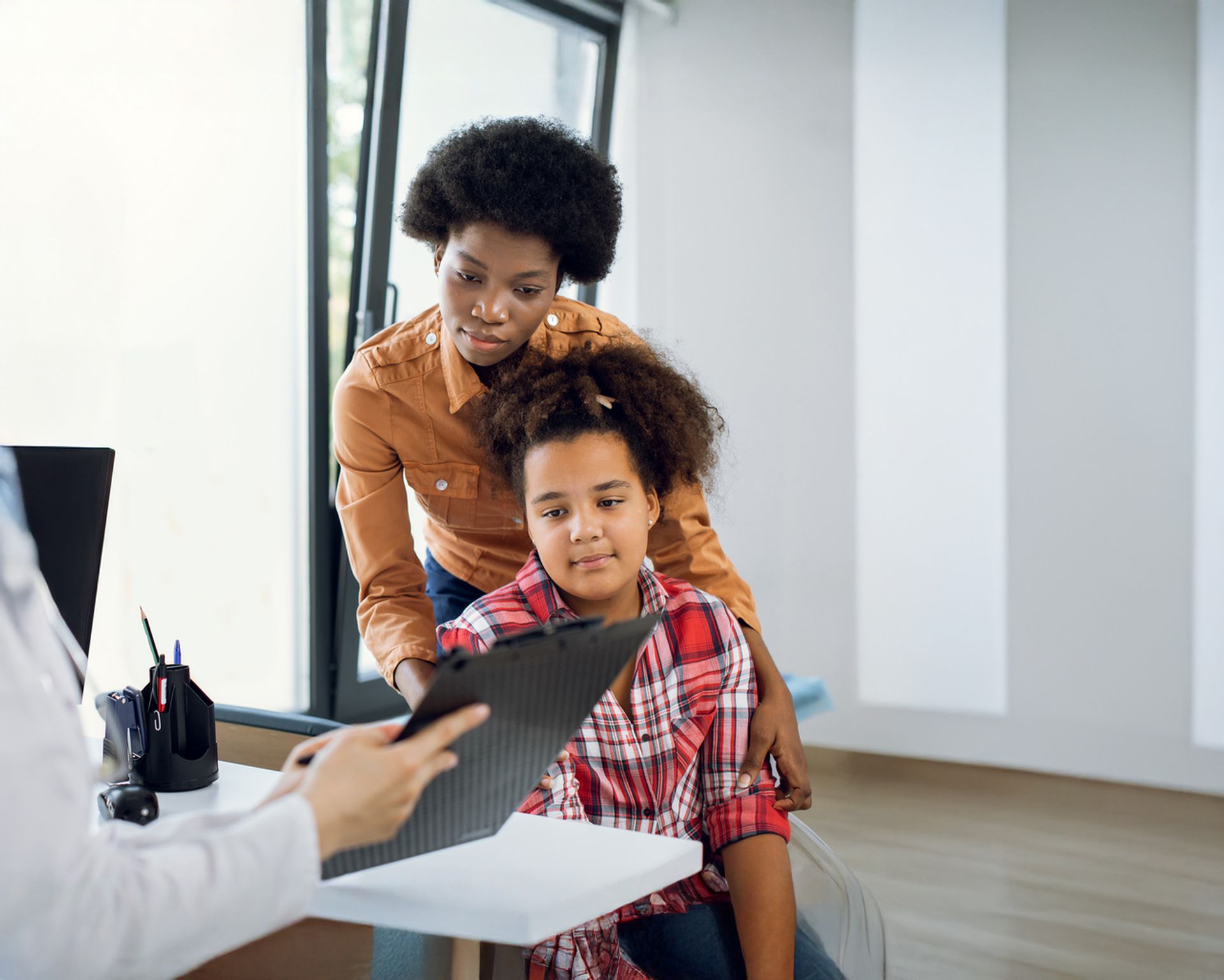 Woman and child at doctor's office; mother comforts girl as doctor holds a clipboard.