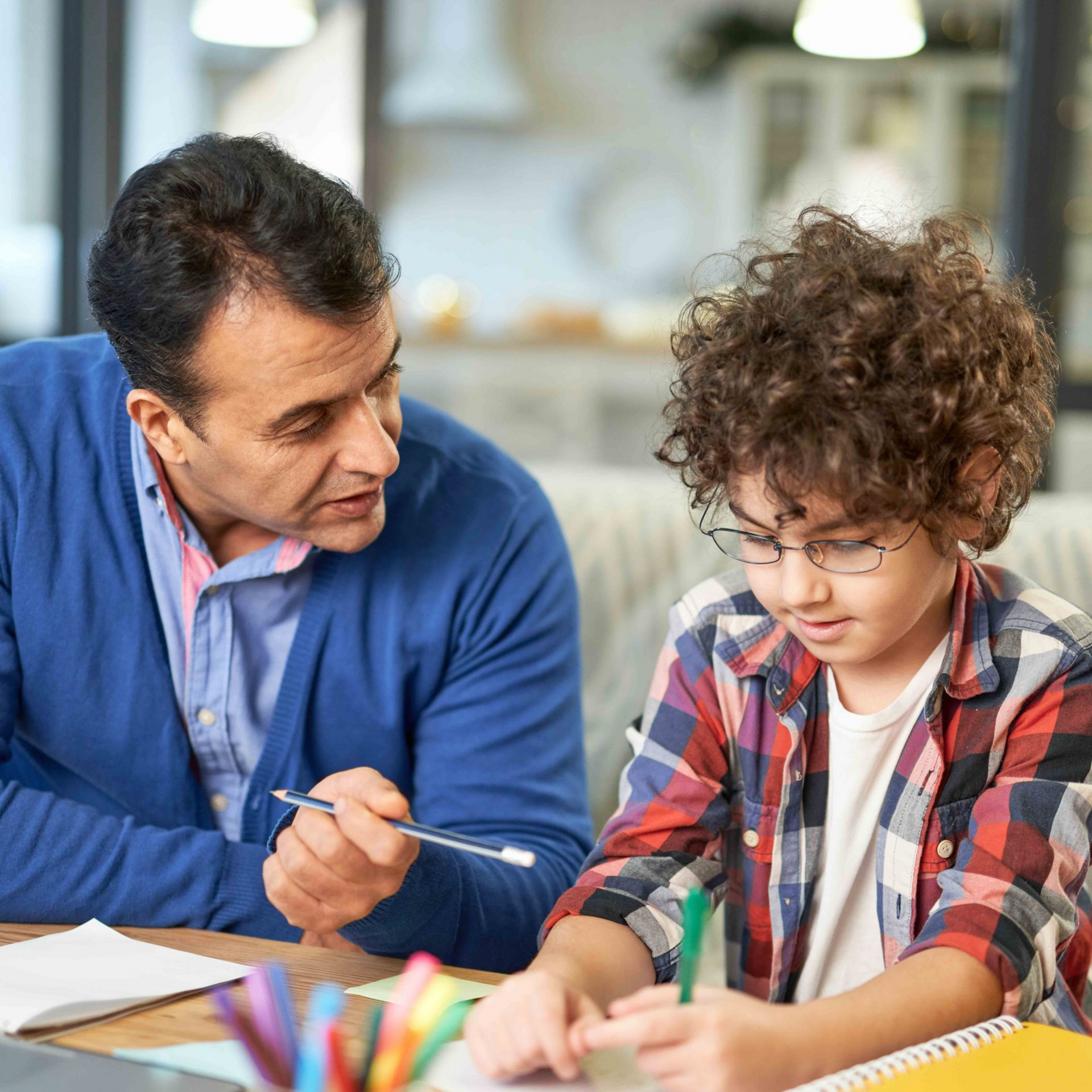 Man helping a child with homework at a desk. The child wears glasses and is writing.