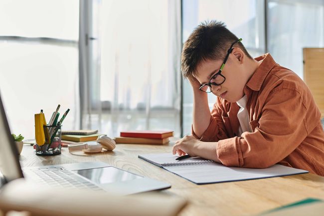Person with Down syndrome wearing glasses, writing in notebook at desk with laptop.