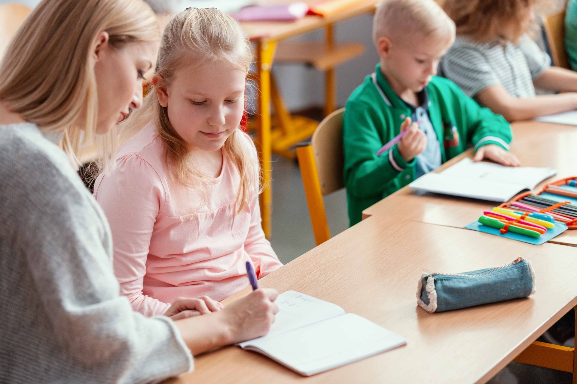 Teacher assisting a student with writing in a classroom; other children work at desks.