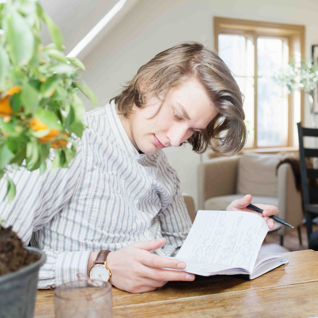 Man with long hair writing in notebook at table, near a potted plant and window.