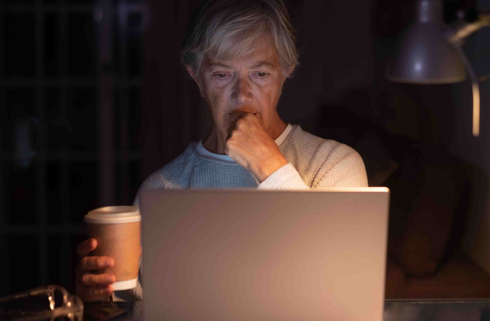 Person focused on a laptop screen, holding coffee, in a dimly lit room, illuminated by a desk lamp.