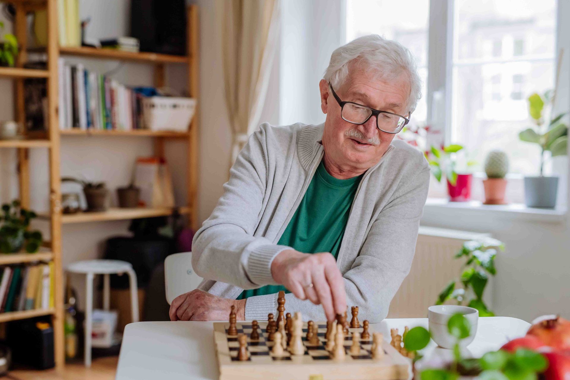 An older person wearing glasses is playing a board game at a table indoors.