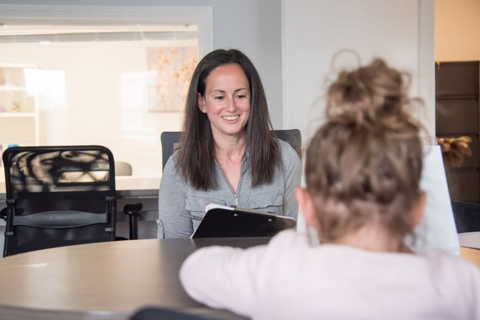 Woman smiles at child across table in an office setting, holding a clipboard.