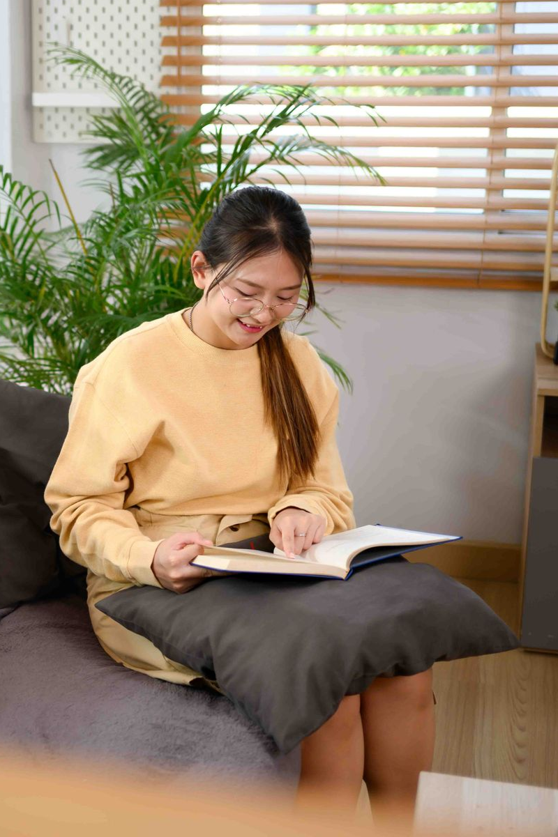 Young person with glasses reading a book, seated on a couch indoors near a window and plants.