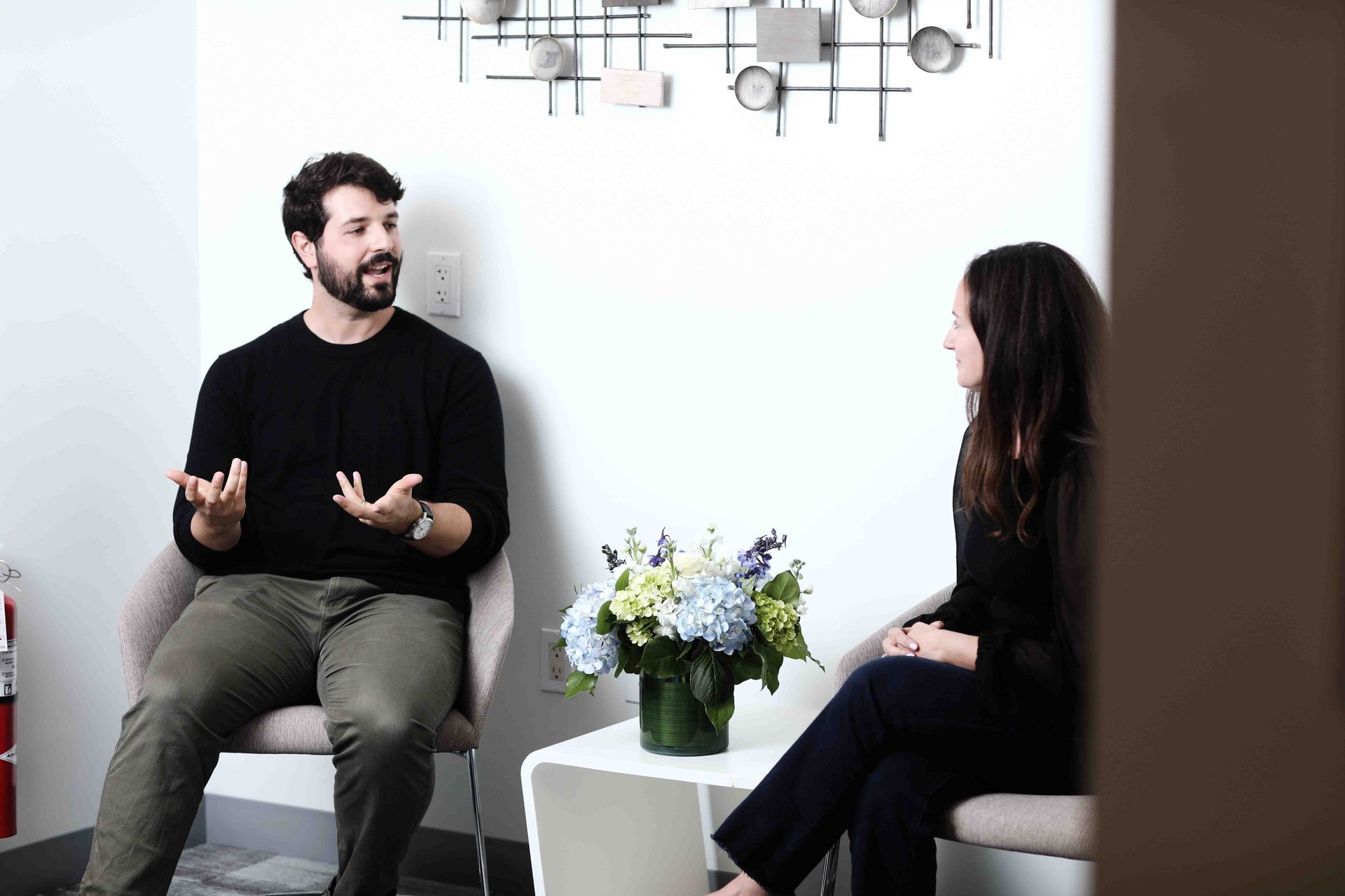 Man gesturing to woman during a conversation in a modern room with floral arrangement.