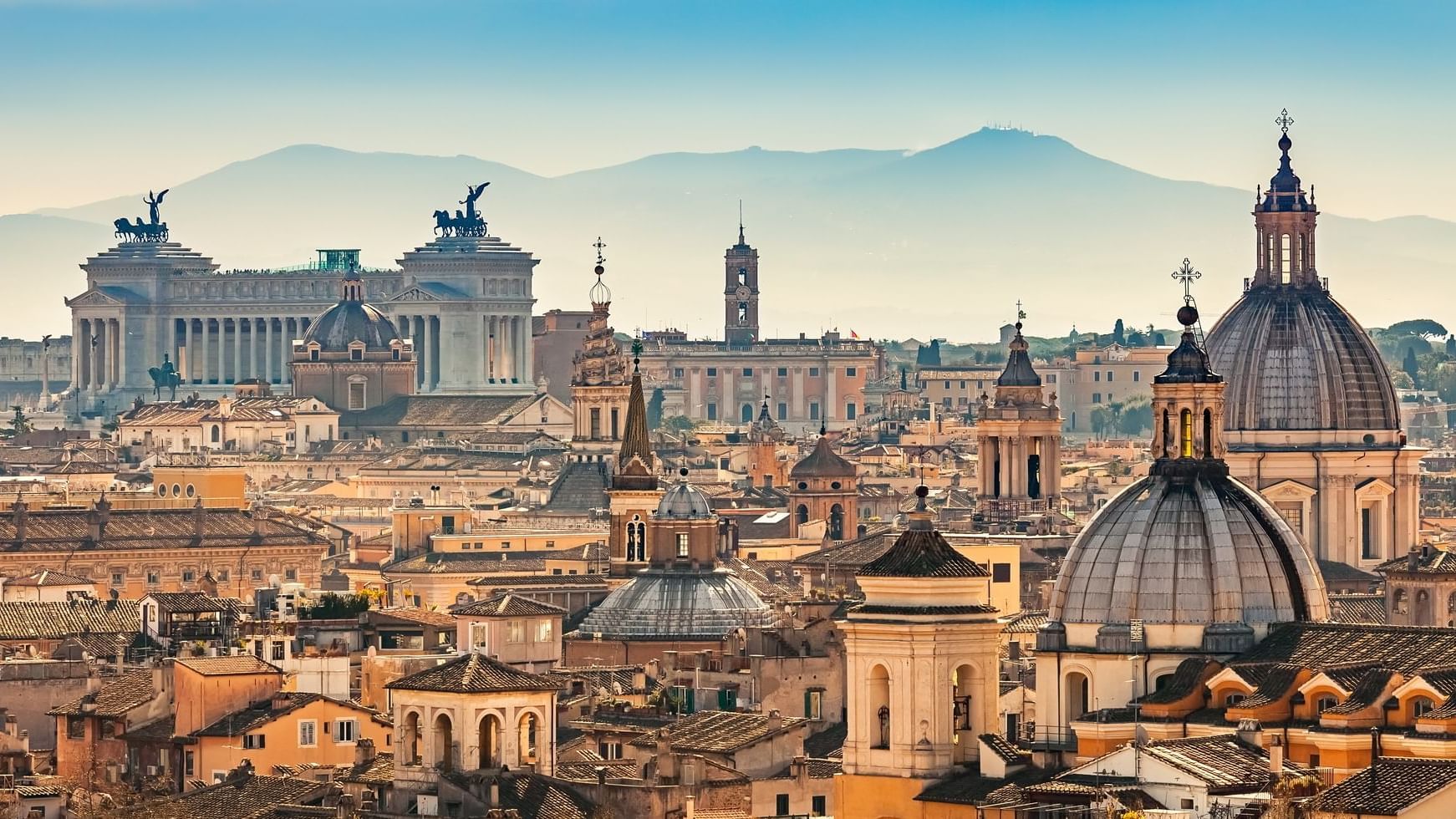 Paesaggio urbano di Roma con cupole, edifici e montagne sotto un cielo nebbioso.