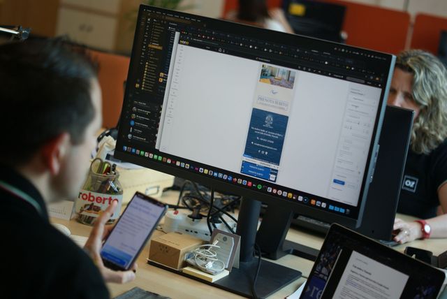 Un uomo usa il telefono e guarda un progetto sullo schermo del computer in ufficio. Un'altra persona lavora al computer.