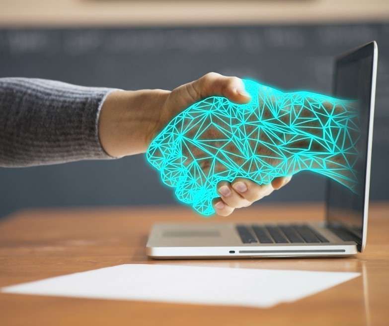 La mano di una persona che stringe una mano digitale blu brillante che emerge dallo schermo di un computer portatile.