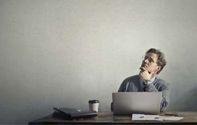 Uomo alla scrivania, con lo sguardo rivolto verso l'alto, con computer portatile, caffè e documenti a portata di mano.