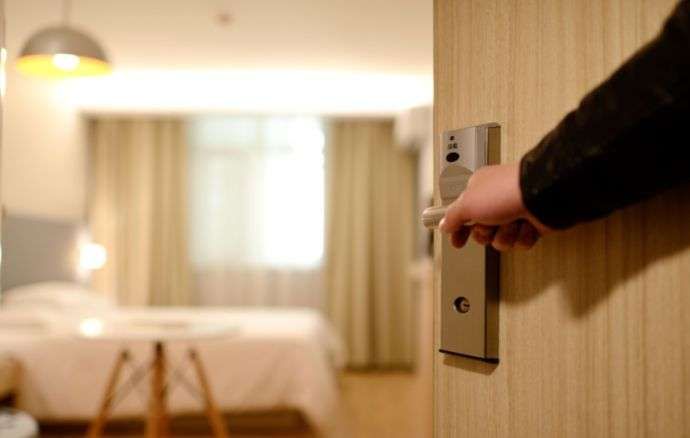 Persona che apre la porta della camera d'albergo; interno sfocato con letto e lampada visibili.