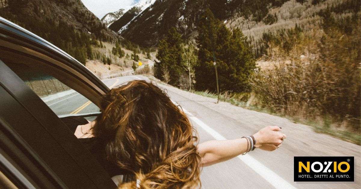 Una donna dai capelli castani si sporge dal finestrino di un'auto su una strada di montagna, con le braccia tese.