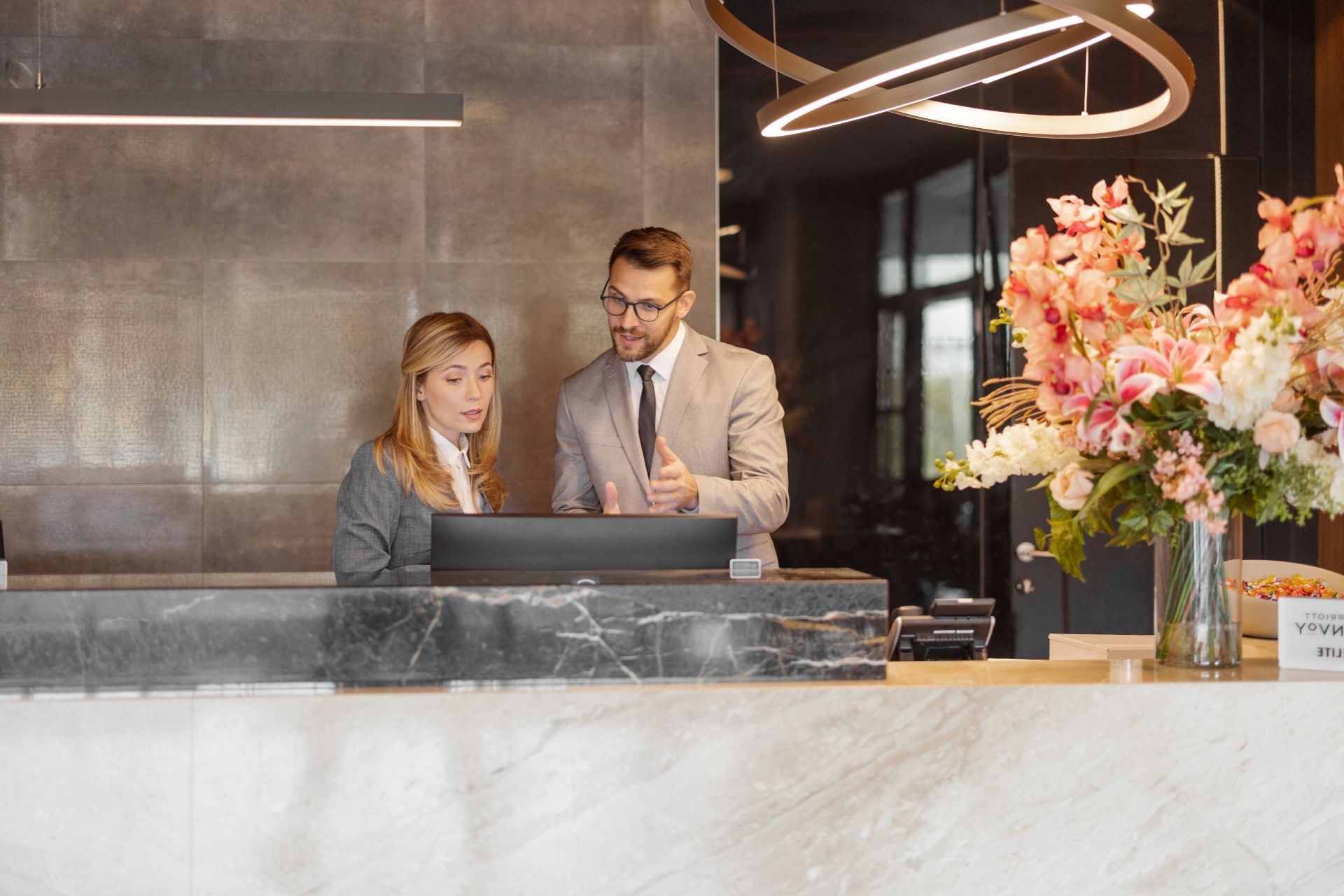 Un uomo e una donna alla reception di un hotel, mentre guardano un computer. Fiori in un vaso, interni moderni.