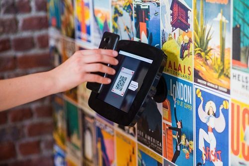 Persona che utilizza uno smartphone per scansionare un codice QR su un tablet montato su una parete di poster di viaggio.