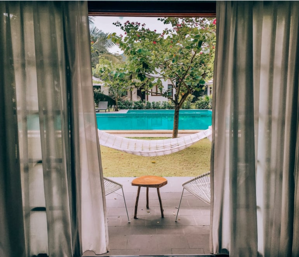 Vista attraverso le porte aperte: piscina, prato con amaca, piccolo sgabello, sedie e alberi.
