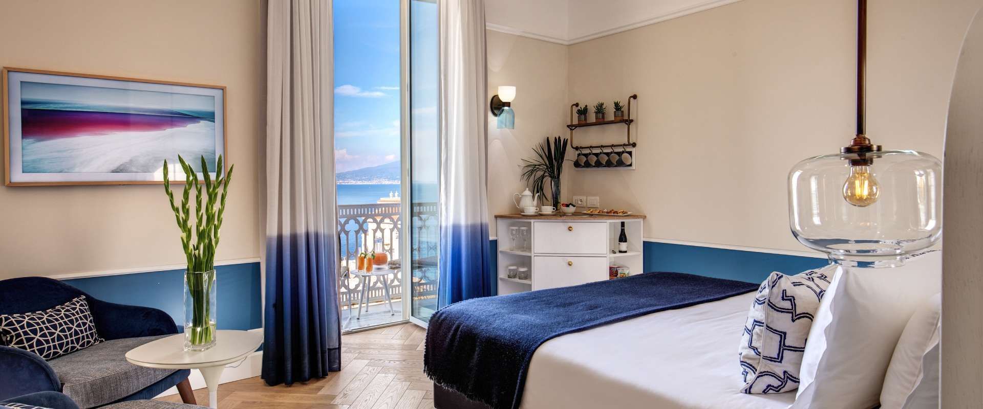 Camera da letto con vista mare, colori blu e bianco, balcone, letto, divano, lampadario in vetro.