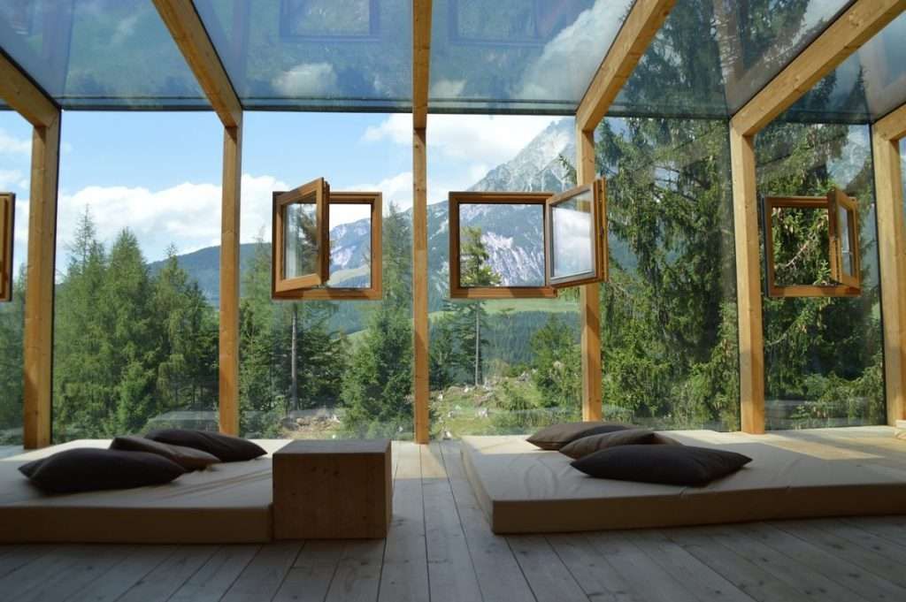 Interno di una struttura in vetro con struttura in legno e finestre aperte, con vista su una foresta di montagna.