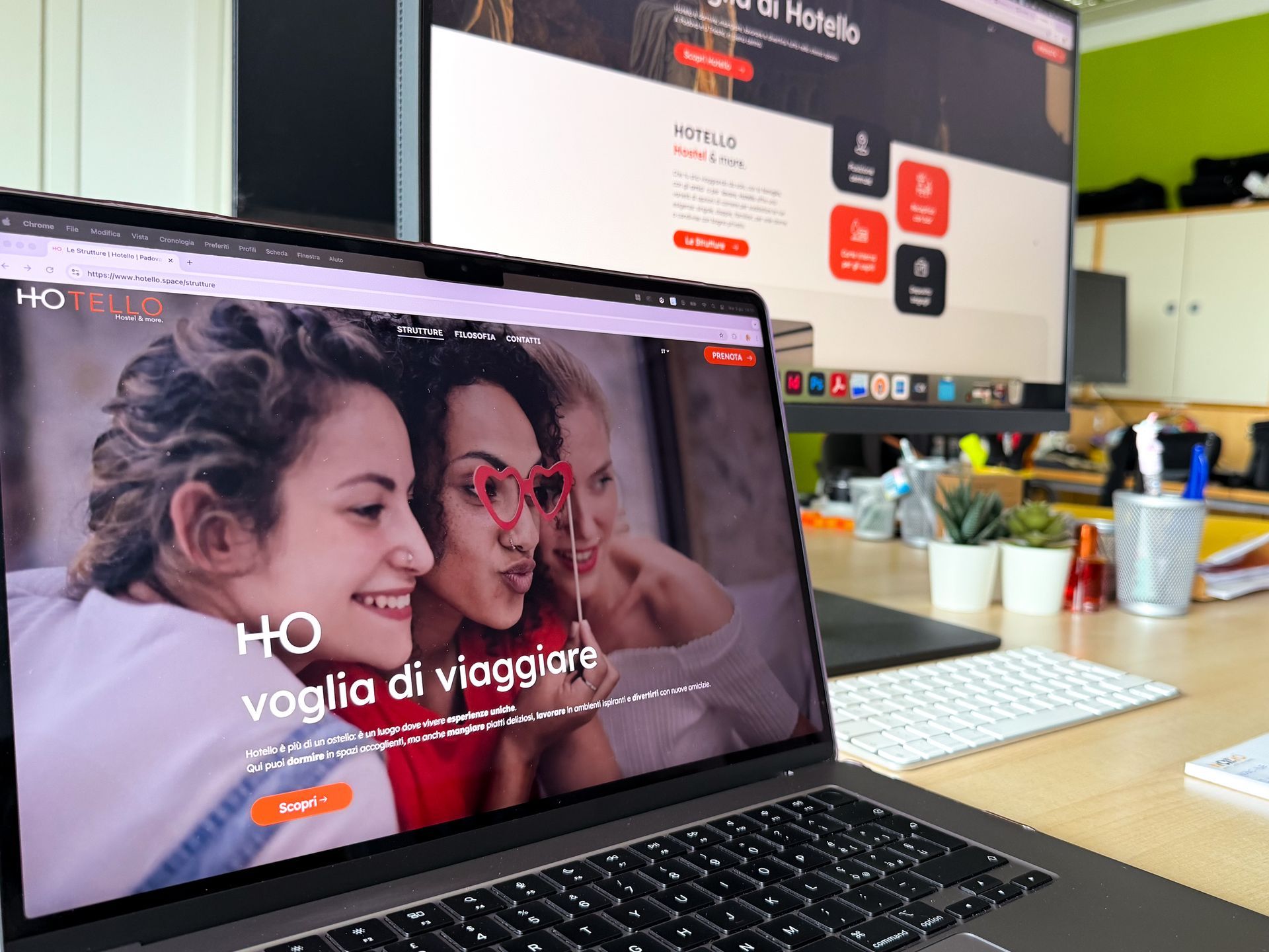 Computer portatile e monitor che visualizzano un sito web, con la foto di tre donne sorridenti. Scrivania.