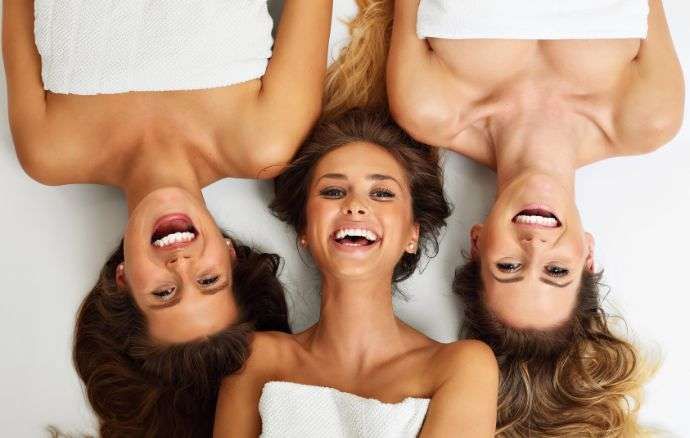 Tre donne sorridenti e ridenti, sdraiate sulla schiena con le teste unite, avvolte in asciugamani bianchi.