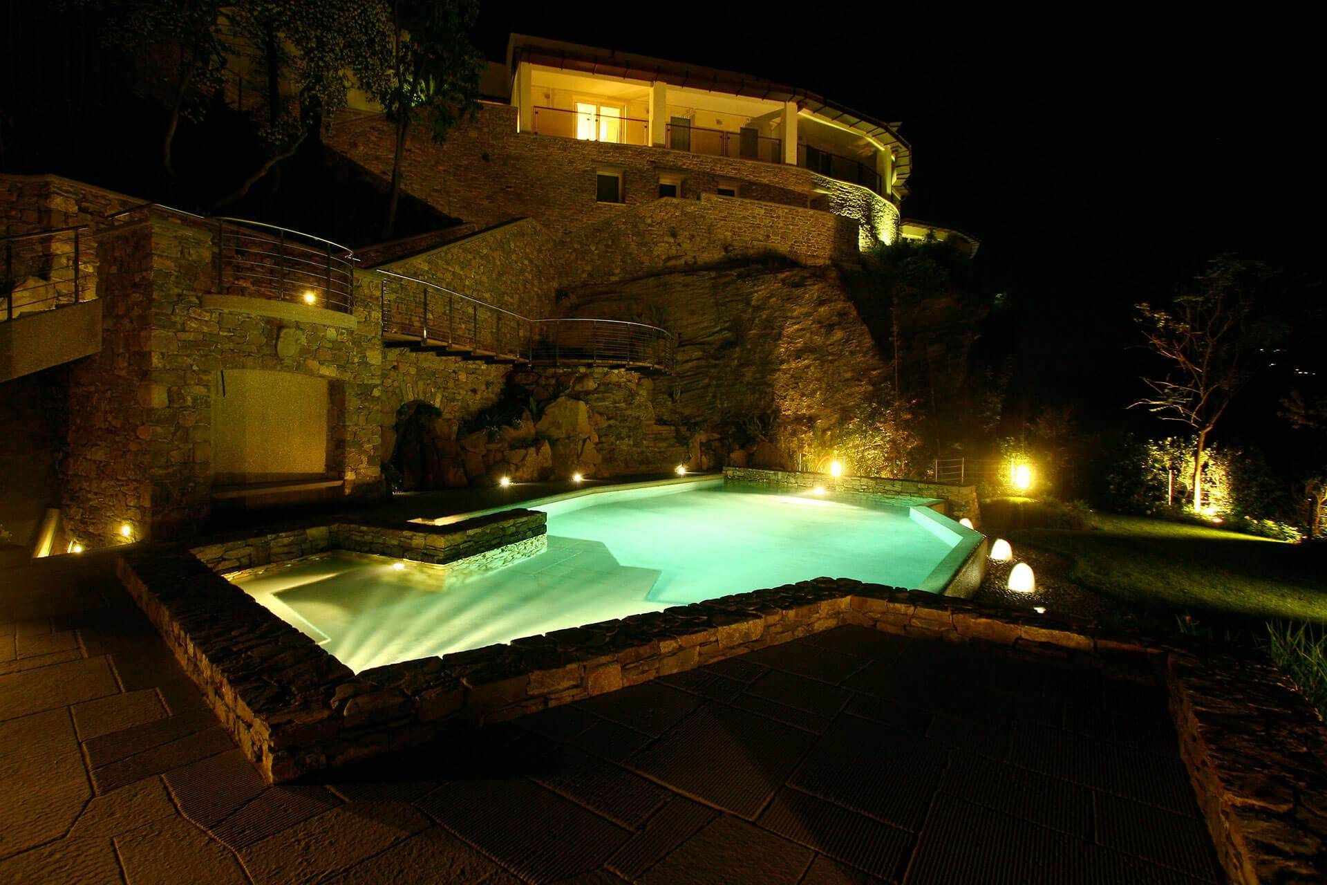 Vista notturna di una lussuosa piscina illuminata da luci verdi, accanto a una casa con muri in pietra su una collina.