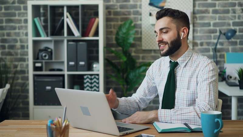 Uomo in camicia e cravatta che usa il computer portatile nel suo ufficio domestico e parla con gli auricolari wireless.