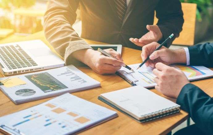 Due persone in giacca e cravatta esaminano grafici finanziari, documenti e un computer portatile su un tavolo.