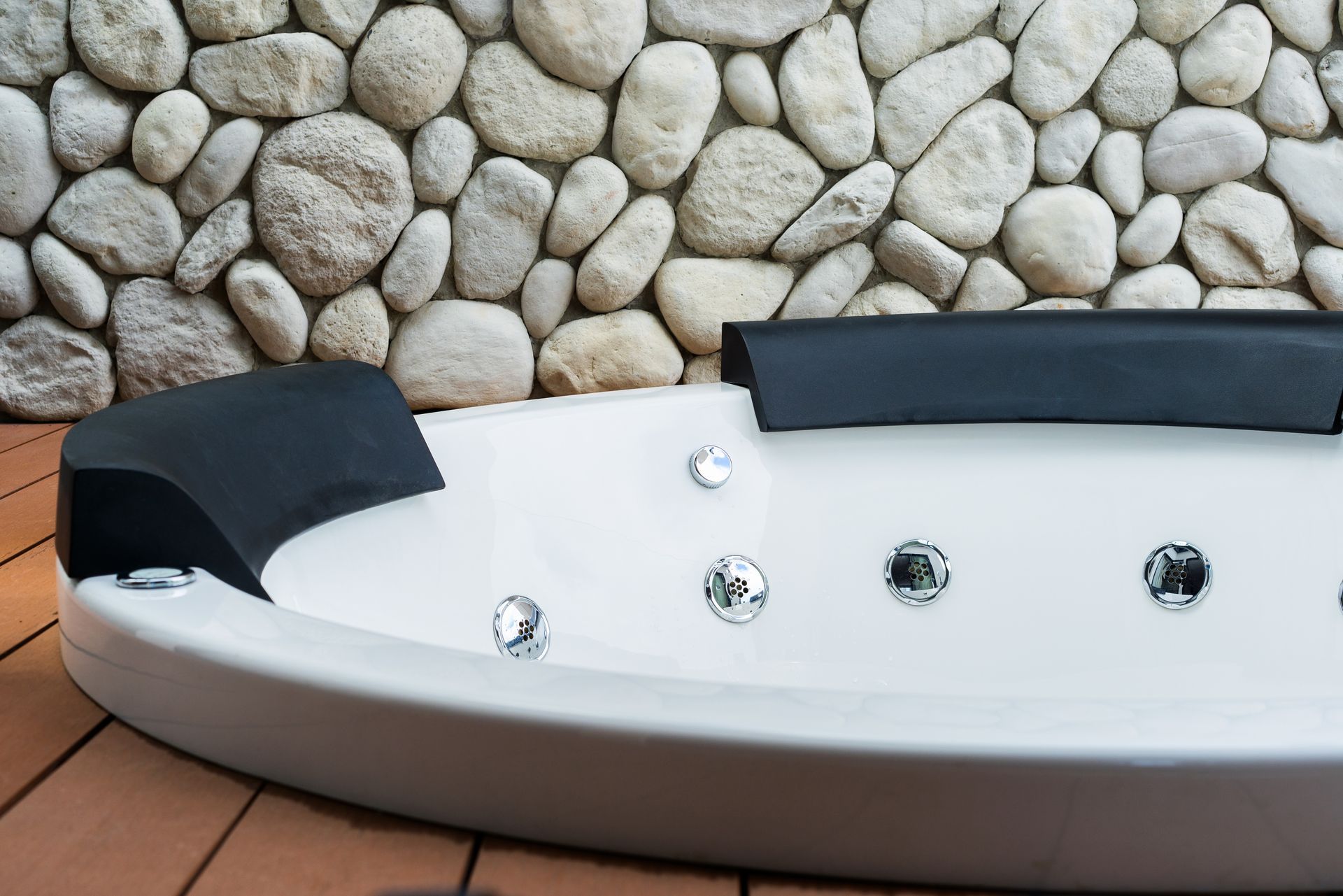 Modern white jacuzzi tub with black headrests against a stone wall and wooden deck
